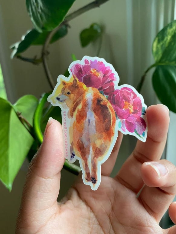 A sticker of a shiba inu and flowers in a hand in front of plants
