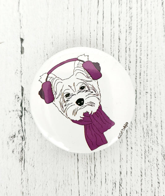 A small white round button pin of a Westie wearing pink earmuffs and a pink scarf