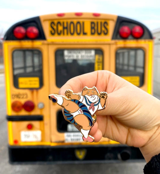 A shiba schoolgirl enamel pin held in a person's hand in front of a yellow schoolbus